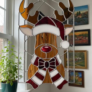 Op de afbeelding: Gebrandschilderd glazen kerstdecoratie van een rendier. Het rendier heeft een rode neus, een kerstmuts en een rood-witte gestreepte sjaal. De geweien zijn amberkleurig en bruin. Het glas is omlijst met zilverkleurig metaal.