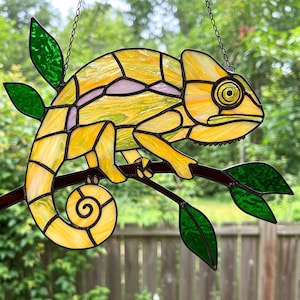 May include: A stained glass chameleon with yellow, green and pink accents perched on a branch with green leaves. The chameleon's tail is curled up.