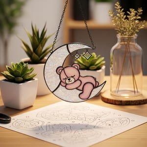 Op de afbeelding: Een gebrandschilderde zonnevanger met een slapende roze teddybeer op een halve maan. De maan heeft een parelmoer afwerking en is versierd met twee hangende sterbedels. De zonnevanger hangt aan een zwarte ketting. Een knutselproject is zichtbaar op de voorgrond.