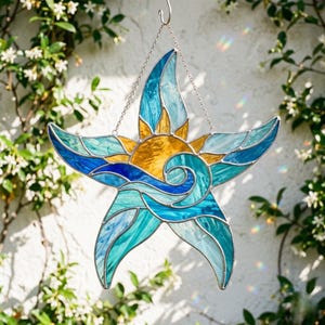 May include: A star-shaped stained glass suncatcher featuring a central yellow sun surrounded by swirling blue and teal ocean waves. The piece is constructed with individual glass segments held together by silver metal solder lines and hangs from a chain.
