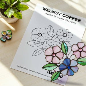K&ouml;nnte beinhalten: Ein schwarz-wei&szlig;es Bleiglasmuster f&uuml;r einen Blumenstrau&szlig; mit nummerierten Abschnitten. Das Muster tr&auml;gt den Titel "Walnut Coffee" und enth&auml;lt den Text "A pattern by Walnut Coffee Pattern". Das Muster wird von einem fertigen Bleiglasst&uuml;ck mit drei Blumen in Rosa, Blau und Lila begleitet.