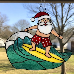 Puede incluir: Un adorno de vidrio tintado que representa a Papá Noel montando una tabla de surf sobre una ola verde. Papá Noel lleva un traje de baño de lunares rojos y blancos y un gorro de Papá Noel rojo.