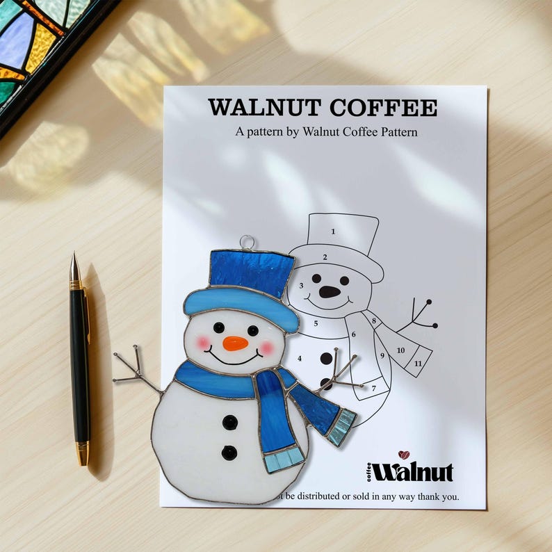May include: A sheet with the text "WALNUT COFFEE" and a stained glass snowman pattern. The snowman has a blue hat, scarf, and accents. A black and gold pen lies to the left. The pattern is by Walnut Coffee Pattern.