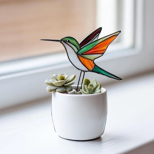 Puede incluir: Un colibrí de vidrio coloreado con alas verdes, naranjas y blancas posado en una maceta de cerámica blanca con dos pequeñas suculentas verdes.