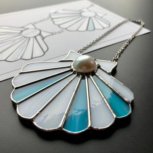 May include: A stained glass seashell pendant with a pearl center, featuring white and shades of blue glass. The pendant hangs from a silver chain. A sketch of the seashell design is visible in the background.