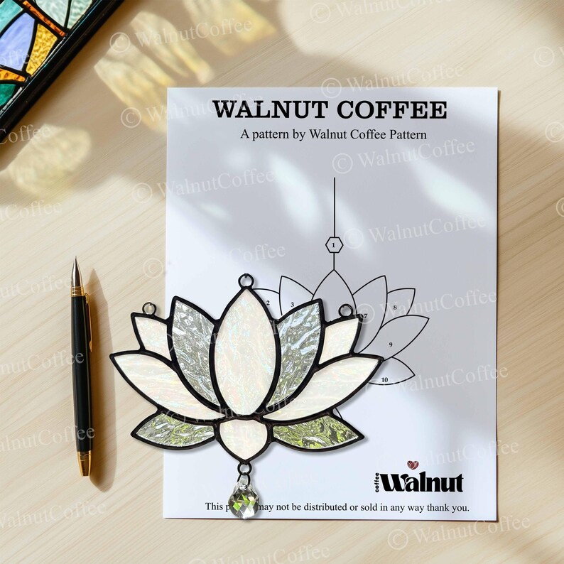 May include: A stained glass lotus flower with white and iridescent petals, green accents, and a clear crystal pendant. The design sits on a pattern sheet with the text "WALNUT COFFEE". A black pen is to the left.