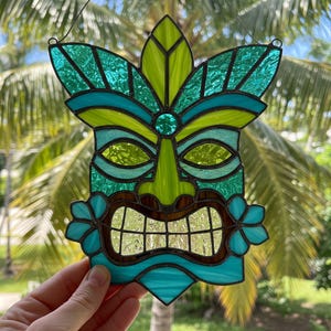 May include: Stained glass tiki mask in shades of teal, green, and brown. The mask features a stylized face with a wide grin, floral accents, and a leaf-like crown. The artwork is designed to hang.
