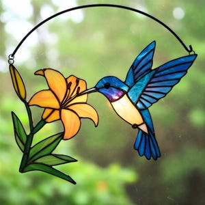 May include: Stained glass hummingbird with blue, purple, and white plumage, feeding from an orange lily. The artwork is framed by a black metal arch, designed for hanging. The vibrant colors and detailed design create a decorative piece.