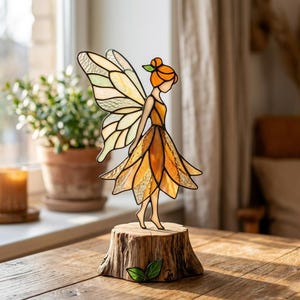 Op de afbeelding: Een gebrandschilderde feeënfiguur met oranje haar en een gele jurk, staand op een houten voet. De fee heeft gedetailleerde vleugels met een mix van geel, groen en helder glas. De basis is een kleine, rustieke boomstronk met groene bladeren.