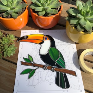 May include: A stained glass toucan bird with a long orange and yellow beak, perched on a brown branch with green leaves. The bird has black and green feathers. The toucan is on a numbered paper with succulents in orange and yellow pots.