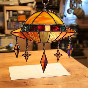 May include: A stained glass UFO ornament with a yellow, orange, red, and green design. The UFO has five dangling stars, two of which are gold, two are purple, and one is red.