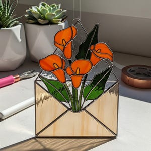 May include: Stained glass art piece featuring orange calla lilies and green leaves arranged in a light-colored envelope shape. The glass has a textured appearance, and the piece is designed to hang. The background includes white pots with plants.