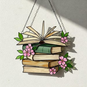 May include: A stained glass window hanging with a stack of books and pink flowers. The top book is open with pages fanned out.