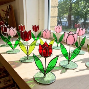 May include: A collection of stained glass tulips in various shades of pink and red. Each tulip is mounted on a green metal stand with leaves.