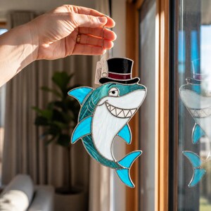May include: A stained glass shark ornament, with a top hat, in teal, white, and blue. The shark has a smiling face and is suspended by a chain. The top hat is black with a burgundy band.