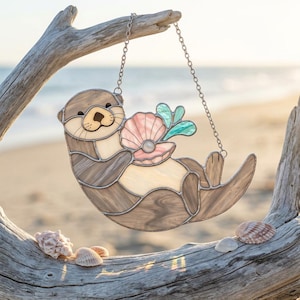 Pode incluir: Um apanhador de sol de vitral em forma de lontra segurando uma concha rosa com uma pérola branca dentro. A lontra é feita de peças de vidro marrom e creme com linhas de solda escuras. Pendura-se por uma corrente de metal prateada nas bordas.