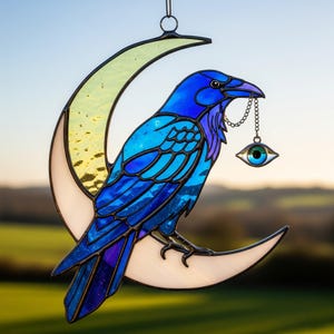 Puede incluir: Vidriera de un cuervo azul y morado posado en una luna creciente. El cuervo sostiene una cadena con un dije de ojo. La luna es blanca y amarilla. Una pieza decorativa para el hogar.