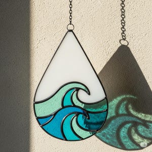 Könnte beinhalten: Ein tropfenförmiger Glas-Suncatcher mit Wellenmuster. Der obere Teil ist weiß, der untere Teil zeigt türkisfarbene und blaue Wellen. Er hängt an einer Kette und wirft einen wellenförmigen Schatten.