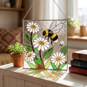 May include: Stained glass panel featuring a bee and daisies. The bee is black and yellow with iridescent wings, hovering near white daisies with yellow centers and green leaves. The panel is square and hangs from a silver chain.