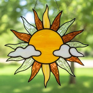Puede incluir: Atrapasoles de vidriera con un sol amarillo brillante con rayos naranjas y amarillos. Dos formas de nubes blancas están debajo del sol. El atrapasoles está diseñado para colgar y tiene un borde negro.