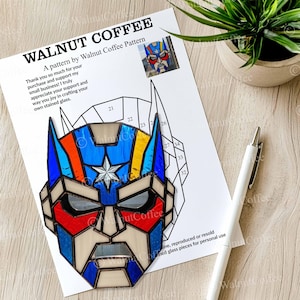 May include: A stained glass pattern by Walnut Coffee, featuring a superhero mask design. The pattern includes a star, blue, red, and yellow accents. A white pen and a small potted plant are also in the image.