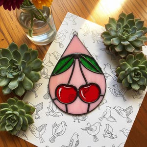 May include: A stained glass cherry pendant with red cherries, green leaves, and a pink background. The pendant is surrounded by black lines and has a metal loop for hanging. The background is a white paper with bird illustrations. Succulents and a vase of flowers are also in the image.