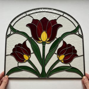 May include: Stained glass panel featuring three tulips in shades of burgundy and yellow, with green stems and leaves. The panel has a semi-circular top and is framed with dark metal. The background is a translucent white.