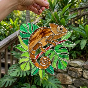 May include: Stained glass chameleon suncatcher. The artwork features a brown and tan chameleon on a red and white striped branch, surrounded by green leaves. It is suspended by a chain and is suitable for hanging.