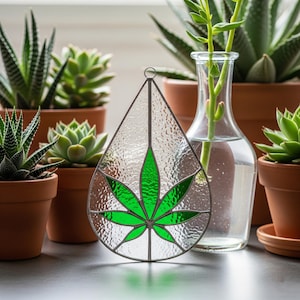 May include: A stained glass teardrop-shaped ornament featuring a green cannabis leaf design. The ornament is surrounded by a clear, textured glass and a silver-colored metal frame. The background includes several potted succulents and a glass vase with a plant.