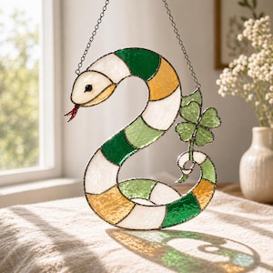 Op de afbeelding: Gebrandschilderde slang zonnevanger met een klavertjevier. De slang is gesegmenteerd met wit, groen en geel glas. Het heeft een rode tong en zwarte ogen. De klaver is groen. De zonnevanger hangt aan een ketting.