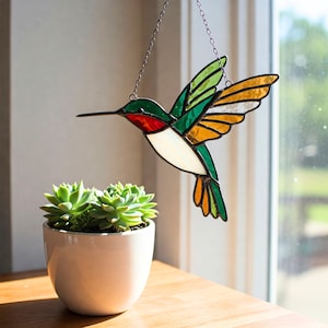 May include: A stained glass hummingbird in flight, suspended by a chain. The bird features green, red, white, and orange glass panels. A small succulent plant in a white pot sits below the hummingbird near a window.