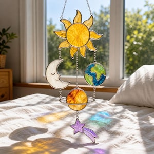 Puede incluir: Un atrapasoles de vidriera con temática celestial que presenta un sol amarillo, una luna creciente con los ojos cerrados, una Tierra azul y verde, un planeta con anillos y una estrella fugaz púrpura. El atrapasoles cuelga de una cadena.