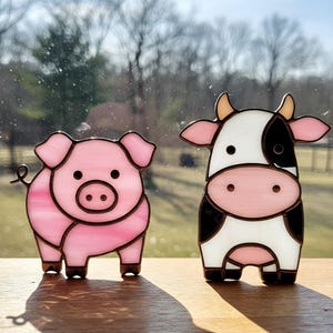 Puede incluir: Dos figuritas de animales de vidrio coloreado, un cerdo rosa y una vaca blanca y negra, sobre una superficie de madera.