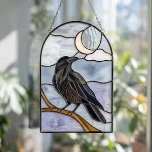 Puede incluir: Un atrapasoles de vidrieras en forma de arco que muestra un cuervo negro posado en una rama dorada sobre un cielo azul pálido y lavanda con una luna creciente blanca y nubes. La pieza está enmarcada en metal oscuro y cuelga de una cadena.