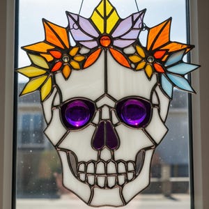 May include: A stained glass skull with a floral crown. The skull has purple eyes and nose, and a white face. The crown is made of yellow, orange, and blue flowers. The piece is framed with dark metal and is hanging in front of a window.