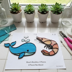 May include: Four stained glass pieces, a blue whale, a pink shrimp, and two smaller pieces, on a white background. The pieces are labelled 1.2 and 1.5. The text "Beach Buddies 4 Pieces" and "4 Pieces Numbeed" is printed below the pieces. The text "Numereana" is printed in the bottom right corner.