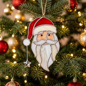 May include: A stained glass Santa Claus ornament featuring a red hat with a white trim and a white pom-pom, a peach-colored face, and a long white beard. A small silver chain hangs from the hat, ending in a clear, faceted glass star charm.