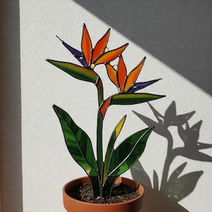 Puede incluir: Una flor de ave del paraíso de vidrio coloreado en una maceta de terracota. La flor es naranja, amarilla y azul. Las hojas son verdes.