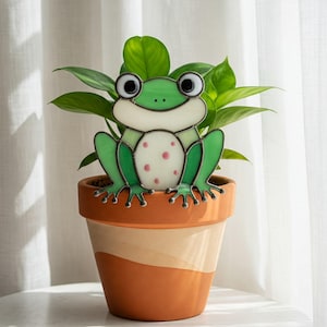 May include: A green stained glass frog with pink spots sits on the rim of a terracotta pot with a plant growing out of it.