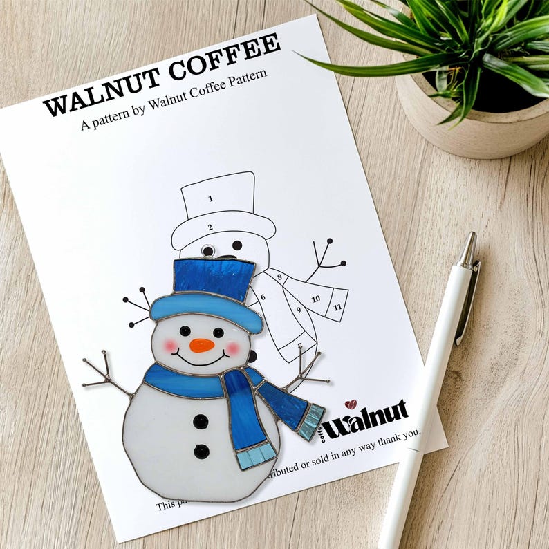 May include: A printed pattern sheet titled "WALNUT COFFEE" featuring a stained glass snowman design. The snowman has a blue hat, scarf, and black button eyes. A white pen and a potted plant are also in the image.