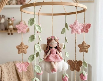 May include: A crochet nursery mobile featuring a central fairy doll with brown hair and a pink dress sitting on a white cloud. The mobile includes hanging elements such as light green leaves, pink and tan stars, butterflies, and pink rose buds.