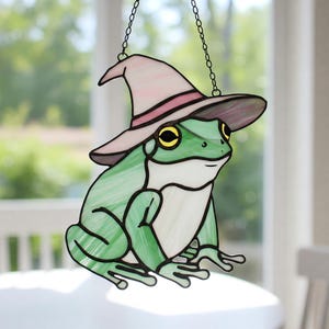 Peut inclure: Une grenouille en verre coloré portant un chapeau de sorcière rose et violet. La grenouille est verte avec des yeux jaunes et est assise sur un fond blanc.