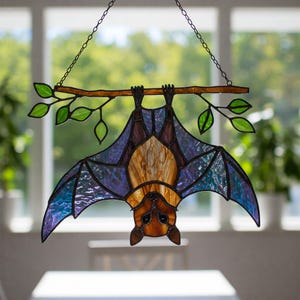 May include: A stained glass bat hanging from a branch with purple, blue, and brown accents. The bat is hanging upside down with its wings spread out.