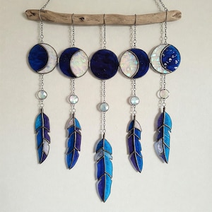 May include: A stained glass moon phase wall hanging with five blue and white moons and five blue and purple feathers hanging from a driftwood branch.