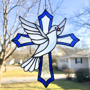 May include: A stained glass window hanging with a blue cross and a white dove. The dove has a red beak and orange eyes.