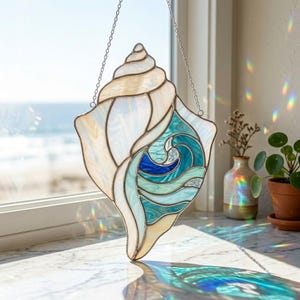 May include: Stained glass seashell suncatcher with a wave design. The shell features iridescent white and cream-colored glass, with a blue and teal wave pattern. It hangs from a silver chain, perfect for coastal decor.