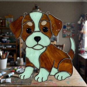 May include: A stained glass puppy dog suncatcher in shades of brown, white, and light green. The puppy has black eyes and a black nose. The glass is outlined with a metal border and has two metal loops for hanging.