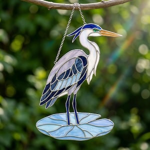 May include: A stained glass suncatcher depicting a heron standing on a lily pad. The bird features shades of blue, white, and lavender glass with a long orange beak and a green eye. It is suspended by a silver metal chain from a branch.