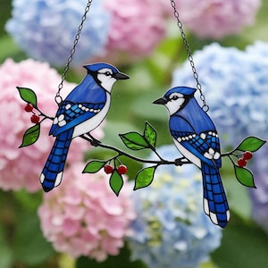以下が含まれることがあります： 緑の葉と赤い実が付いた枝に止まっている2羽の青い鳥を描いたステンドグラスのアート作品。鳥は青と白です。ピンクと青のアジサイを背景に、チェーンで吊るされています。
