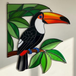 Peut inclure: Vitrail représentant un toucan perché sur une branche avec des feuilles vertes. L'oiseau a un grand bec orange et jaune, des plumes noires, une poitrine blanche et des yeux bleus. Les feuilles sont de différentes nuances de vert.